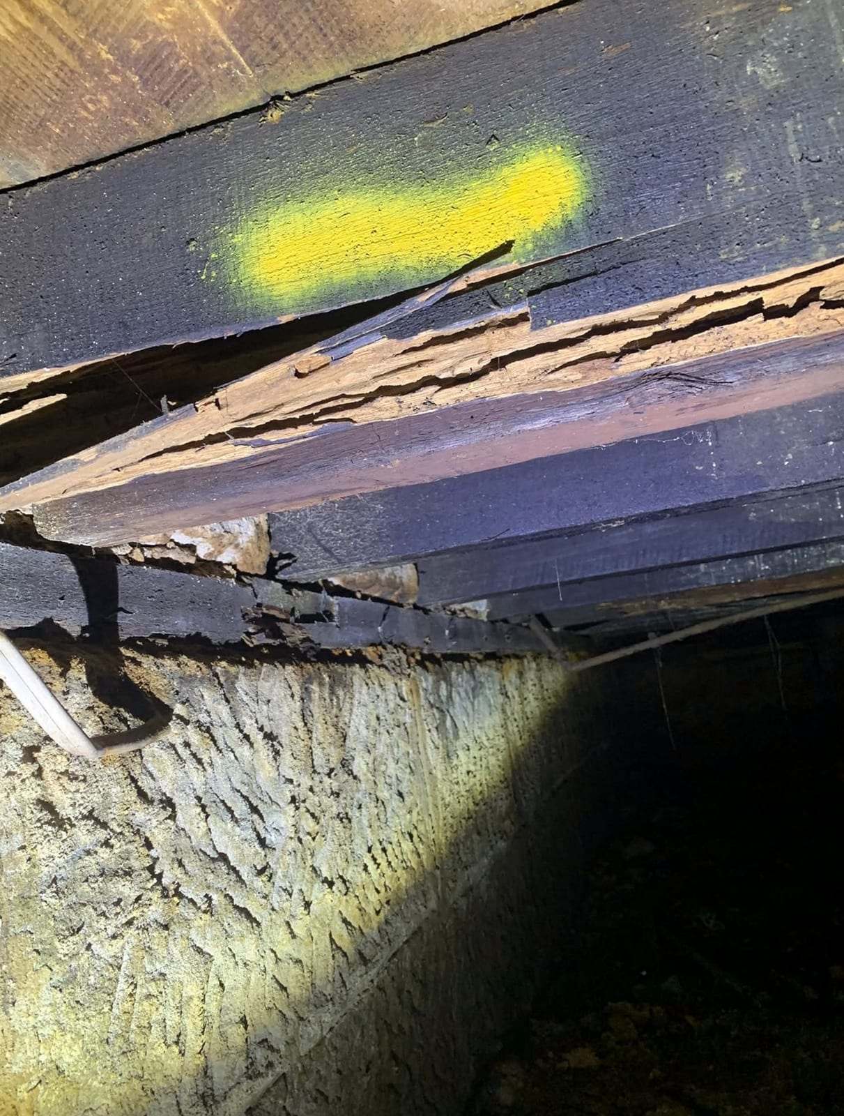 Termite-damaged wooden bearer under a floor, marked with yellow paint and lit by a flashlight in a crawl space